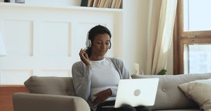 Happy young african woman wear wireless headphones make video call online chat look at laptop screen sit on sofa, smiling girl talk to webcam communicate by internet computer conference app at home