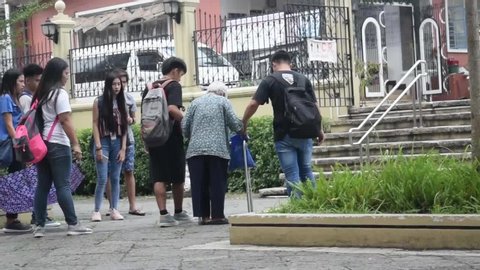 San Pablo City, Laguna, Philippines - October 18, 2019: Old sick woman walking with the aid of a walker aided by good young people to cross the church yard