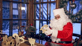 Santa Claus is sitting in specially decorated room at the table, holding wooden dear near. Christmas spirit concept close up video. Christmas eve. New Year is coming. Presents in Santa Claus's hands.