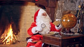 Santa Claus is sitting in specially decorated room at the table, holding a magnifying glass in hands. Feast fireplace in the background. Christmas spirit concept close up video. Christmas eve.