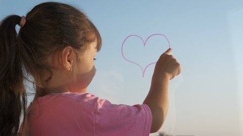 The child on the window draws a heart. Little girl traces a finger drawn heart over glass.