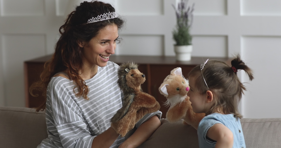 Playful happy young mum babysitter wearing crown holding puppet toys on hand talking laughing playing with cute little preschool daughter telling fairy tale having fun together sit on sofa at home Royalty-Free Stock Footage #1039337834