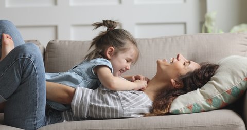 Happy carefree mother and cute small child daughter tickling laughing having fun lying on sofa and playing, smiling mum enjoying bonding cuddling hugging spending time with little funny kid at home