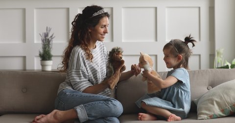 Happy family young mom babysitter or nanny wearing crown holding toys having fun with cute small child daughter sit on sofa, mother queen and little kid girl bonding playing puppets together at home