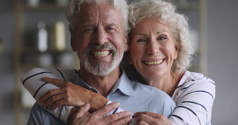 Cheerful old elderly adult family couple hugging laughing bonding looking at camera, smiling healthy senior retired grandparents husband and wife happy faces embracing at home, close up portrait