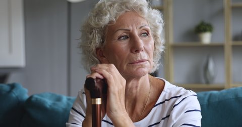 Depressed thoughtful elderly adult woman holding walking cane stick sit alone on sofa, pensive sad old senior grandma feeling lonely suffer from geriatric health problems concept, close up view