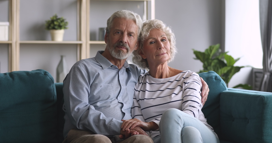 Serious happy senior elderly family couple bonding looking at camera hold hands sit on sofa, smiling old adult grandpa husband embrace grandma wife on couch at home, retired grandparents portrait Royalty-Free Stock Footage #1039337987