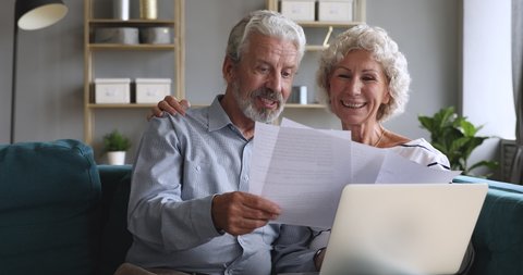 Happy old couple talking checking papers holding domestic bills and laptop computer, satisfied bank clients senior grandparents customers planning budget investment loan insurance payment at home