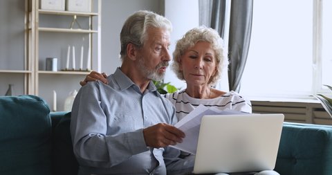 Serious old retired couple holding papers and laptop checking domestic bills planning finances expenses pay online, senior grandparents manage household payments doing paperwork sit on sofa at home