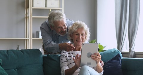 Smiling senior old couple talking using digital tablet computer modern tech at home on weekend lifestyle, happy elderly grandmother and grandfather doing internet shopping choose sale offers together