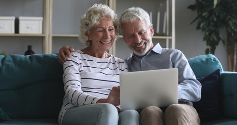 Happy old retired couple talking laughing using laptop doing internet shopping choose sale offers together, senior elderly family grandparents relaxing looking at computer screen sit on sofa at home