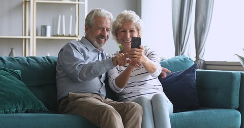 Happy senior elderly couple hold smart phone having fun with mobile app tech take selfie on cell together, cheerful old retired grandparents laugh look at cellphone screen relax sit on sofa at home