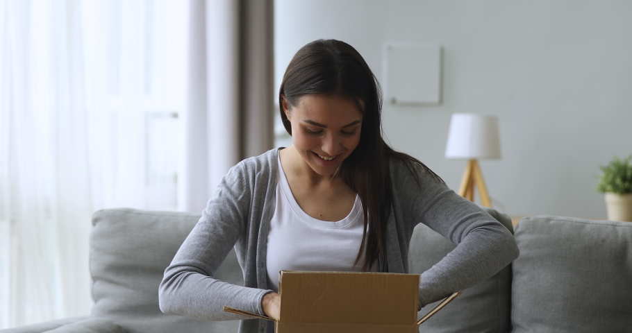 Satisfied female customer open online order shopping courier delivery parcel sit on sofa, happy young woman holding cardboard box unpack receive good purchase by post mail shipping at home concept Royalty-Free Stock Footage #1039338107