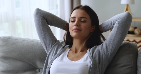 Serene attractive young woman resting on couch taking deep breath of fresh air holding hands behind head, healthy calm lady relaxing on comfortable sofa napping feel stress free at home lounge alone