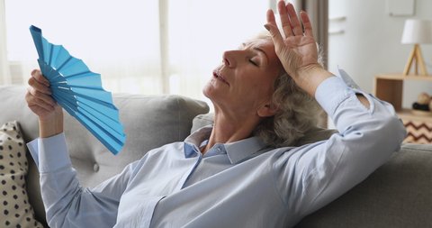 Overheated exhausted old woman feel hot wave fan annoyed with high temperature sit on sofa, tired senior grandma sweating suffer from climax summer heat problem, no air conditioner at home concept