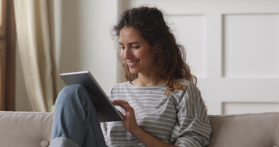 Happy young woman relaxing at home sit on sofa holding digital tablet enjoying surfing internet study work shopping online using social media apps or playing game on modern tech device at home Royalty-Free Stock Footage #1039338188