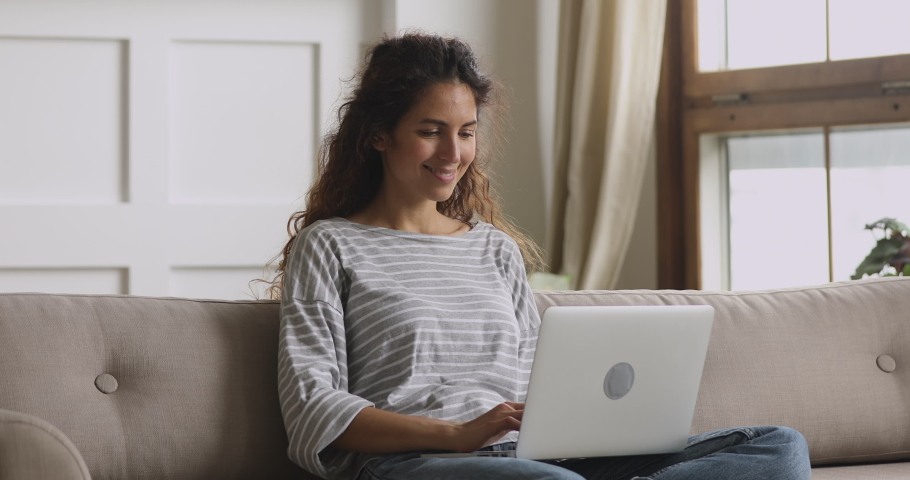 Smiling young woman sitting on couch using laptop notebook looking at screen typing message, happy lady chatting on computer browsing surfing internet social media studying or working online at home | Shutterstock HD Video #1039338191