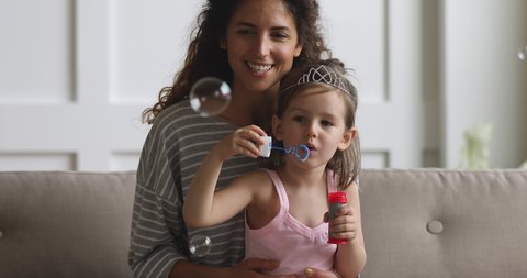 Cute funny little kid girl wear crown playing with happy young mother sitting on sofa, smiling mom babysitter nanny having fun with small preschool child daughter blowing soap bubbles at home