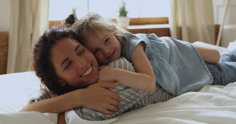 Affectionate family cute adorable funny little kid daughter embrace kiss young mom lying relaxing on bed, happy loving mother and small child girl enjoy bonding hugging having fun playing in bedroom