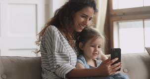 Cute funny little kid girl having fun with mom enjoy using modern gadget smart phone looking at mobile screen laughing making conference call in app, watching social media video sit on couch at home