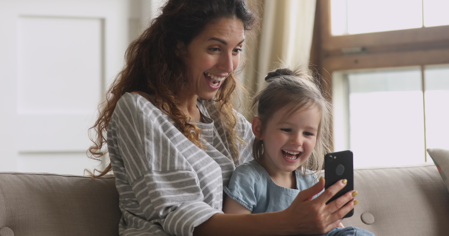 Cute funny little kid girl having fun with mom enjoy using modern gadget smart phone looking at mobile screen laughing making conference call in app, watching social media video sit on couch at home
