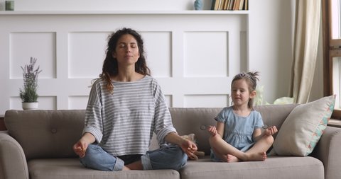 Funny cute little kid daughter meditating in lotus pose laughing with young mom at home, happy healthy family mother and small child girl having fun doing yoga exercise relaxing together sit on couch