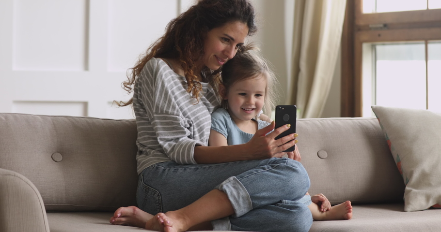 Happy family mum and cute little kid daughter laugh use smart phone funny face mask app look at cell screen, mother with child take selfie watch cartoons make video call on mobile sit on sofa at home | Shutterstock HD Video #1039338266