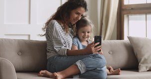 Happy family mum and cute little kid daughter laugh use smart phone funny face mask app look at cell screen, mother with child take selfie watch cartoons make video call on mobile sit on sofa at home