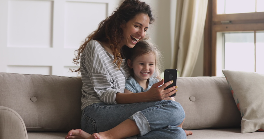 Happy family mum and cute little kid daughter laugh use smart phone funny face mask app look at cell screen, mother with child take selfie watch cartoons make video call on mobile sit on sofa at home Royalty-Free Stock Footage #1039338266
