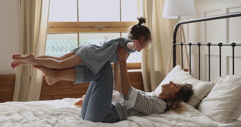 Happy healthy mother holding lifting cute little preschool child daughter up playing plane on bed at home, funny kid girl fly practice acro pair yoga bonding having fun with mum together in bedroom