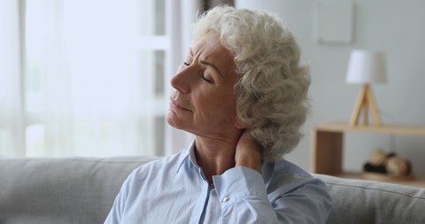 Unhappy tired senior old woman rubbing neck feeling pain sit on couch at home, upset elderly grandma massaging stiff muscles suffering from osteoarthritis osteochondrosis fibromyalgia ache concept