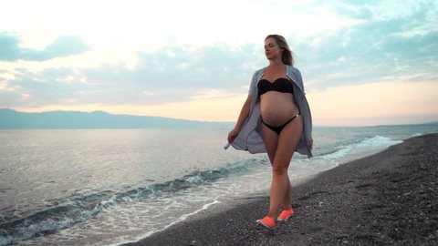Beautiful thinking pregnant woman in swinsuit and long t-shirt walking along the sea beach in amazing pink and blue sunset. Slim pregnant woman's body outdoors. Happy mother relaxing at the sea