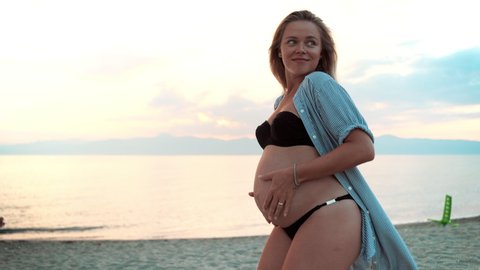 Beautiful woman's pregnancy at the sea sunset background. Happy woman in expectation caressing and embracing lovely her tiny belly with her hands. Healthy pregnancy of happy mother. Pregnant tummy