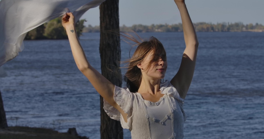 Portrait of a beautiful Caucasian woman in light dress standing on the shore with closed eyes holding scarf in raised hands. Attractive girl enjoying wind blow from the sea.