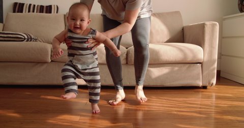 baby first step Slow motion of 6 months baby learning to walk at home mother holding baby arm walking on the floor indoor with sunlight coming family with baby lifestyles