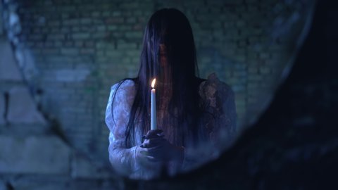 Scary woman with burning candle near mirror, making satanic ritual at night