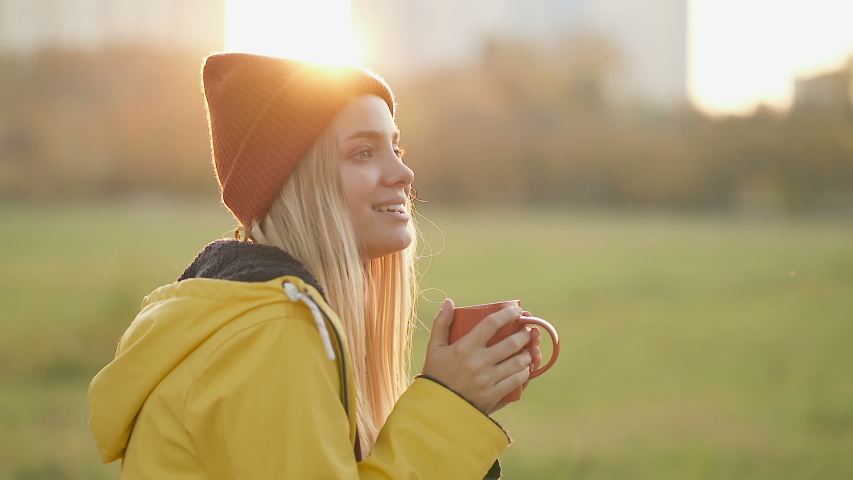 Portrait of happy woman drinking hot tea (or coffee) outdoors, in the park.  Autumn season, sunset time. Slowmotion.