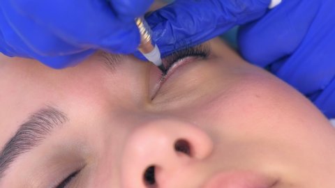 Cosmetologist making permanent eyeliner makeup to woman using tattoo machine, eyes closeup view. Beauty procedure for young girl in beauty clinic salon, hands closeup.