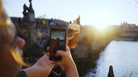 Back view woman enjoying buns against city sights. Girl standing on embankment of Vltava river and taking photo on phone local sweets trdelnik or Trdlo in backlit at blurred background Charles bridge 