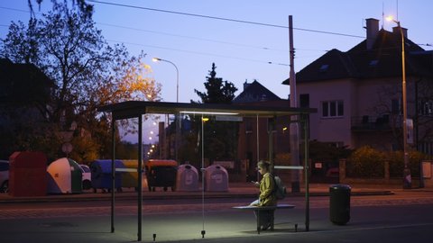Illuminated bus stop in residential area on the outskirts of the city at night. Young woman waiting for public transport inside modern transparent shelter, walking and messaging via smartphone 