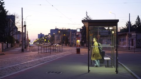 Illuminated bus stop in residential area on the outskirts of the city at night. View through modern transparent shelter lonely young girl waiting public transport, coming to approaching tram