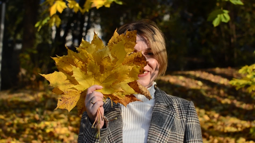 Cute young cheerful and happy girl hides her face behind a yellow maple leaf in autumn park