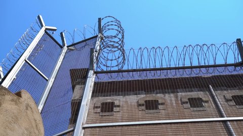 wall of the prison building with small windows with bars and a high fence with barbed wire.