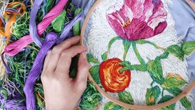 Female hands with needle above composition of embroidery hoop, threads on background of blue embroidered fabric with colorful pattern. Flat lay, top view. Traditional hobby. Preparing for needlework