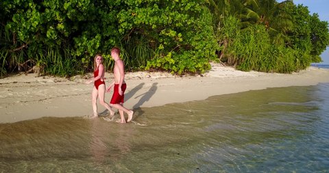 Guy and girl in red swimsuit walking along coastline on shallow waters towards sunbeds in Mentawai Islands