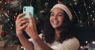 Pretty Young Girl in Santas Hat Sitting under Decorated Christmas Tree and Video Chatting using her Smartphone, Laughing Showing Thumb up Blowing a Kiss