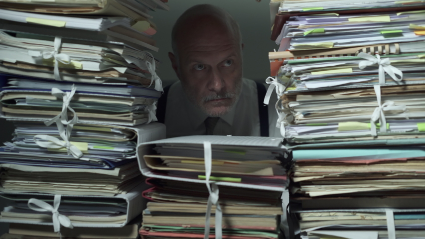Office clerk working behind a wall of paperwork: he is peeking from a hole and stamping a document, bureaucracy concept | Shutterstock HD Video #1039418756
