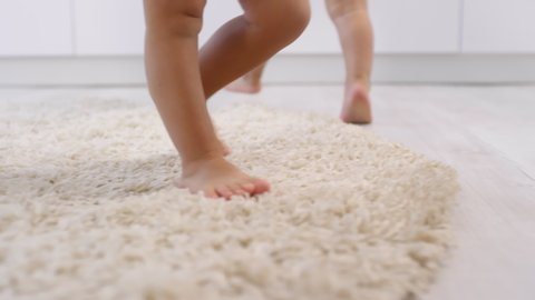 Tracking close-up shot of unrecognizable toddler with bare legs being put down on soft fluffy beige carpet and making wobbly first steps, and twin sibling learning to walk beside