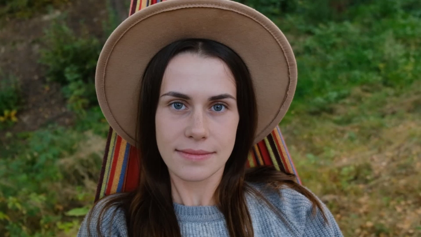 close up portrait of a pretty and beautiful young girl lying on a hammock and looks into camera, smiling happy, enjoying independent lifestyle, dressed grey sweater and hat on autumn day, slow motion