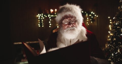Joyful authentic santa clause flipping through pages of red covered book, with fireplace and christmas tree on background - christmas spirit concept close up 4k footage